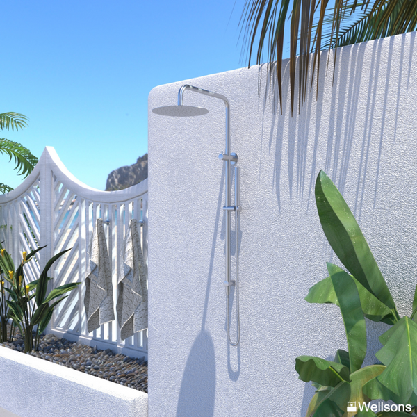 A wall-mounted oval shower head with a chrome slide bar and hose, installed on a white textured exterior wall with palm leaves and tropical plants in a sunny outdoor setting. Wellsons logo is visible in the bottom right corner.